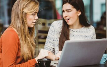 Two young women sit at a table with a laptop and coffee. One looks surprised or shocked while the other appears to explain something, both focusing on the laptop screen.