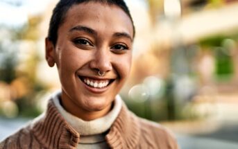 A person with short hair and a nose ring smiles outdoors, wearing a beige turtleneck and a brown textured sweater. The background is blurred with natural light and soft colors.