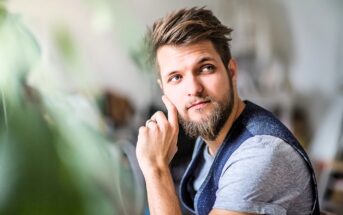 A young man with a beard and mustache, wearing a gray t-shirt and dark vest, sits indoors with his hand resting on his cheek, looking thoughtfully to the side. Green foliage is blurred in the foreground.