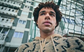 A young man with curly hair wearing a patterned hoodie stands in front of a tall apartment building, looking thoughtful. A green chain-link fence is visible beside him. The photo is taken from a low angle.