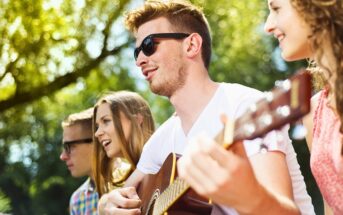 Four young adults sit outdoors on a sunny day. One person plays an acoustic guitar while the others smile and sing along. All are wearing casual clothes, and two of them wear sunglasses. Trees and greenery are in the background.