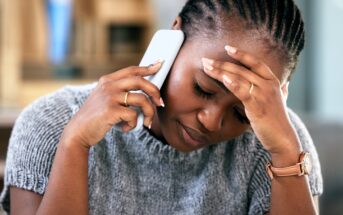 A woman with braided hair looks stressed while talking on the phone, holding her forehead with one hand and her phone with the other. She wears a gray sweater and a watch, and sits indoors.