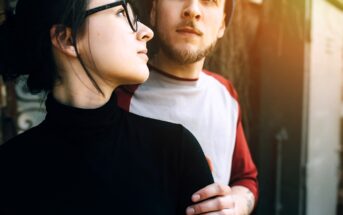 A woman in glasses and a black turtleneck stands closely with a man in a white and red shirt, gently holding his arm. Both look thoughtfully into the distance, illuminated by soft sunlight.