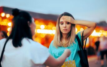 A woman touches her forehead and looks unwell while another person hands her a shopping bag at an outdoor market with blurred lights in the background.