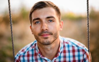 A young man with short brown hair and blue eyes, wearing a red, blue, and white plaid shirt, sits on a swing outdoors, looking directly at the camera, with a blurred natural background.