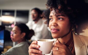 A woman with curly hair holds a white mug and looks thoughtfully ahead, while two colleagues work at computers in the blurred background of an office setting.