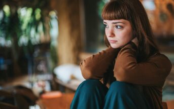 A young woman with straight brown hair and bangs sits indoors, hugging her knees and looking pensively to the side. She wears a brown top and green pants. The background is softly blurred with warm light and plants.