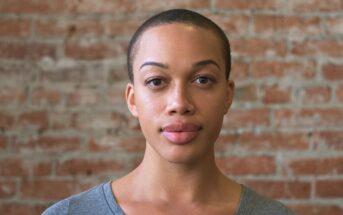 A person with short hair and a gray shirt stands in front of a brick wall, looking directly at the camera with a neutral expression.