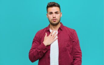 A young man with short dark hair, wearing a maroon button-up shirt over a white t-shirt, stands against a teal background, looking surprised and touching his chest with one hand.