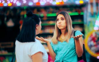 Two women are talking outdoors; one woman with long hair shrugs with her hands up and cheeks puffed out, appearing confused or unsure, while the other woman with dark hair listens. Colorful lights are blurred in the background.