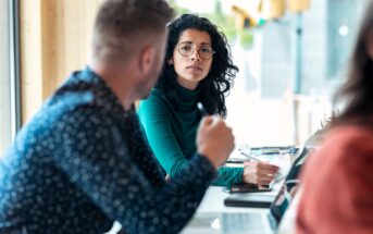 A woman with curly hair and glasses sits at a table, looking attentively at a man who is speaking. They appear to be in a discussion, possibly in a casual office or café setting, with notebooks and laptops on the table.