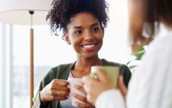 Two women sit together indoors, holding mugs and smiling warmly at each other, suggesting a friendly conversation in a cozy, well-lit room with a lamp and plants in the background.