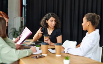 Four women sit at a table with coffee cups, notebooks, and a small plant, having a discussion. One woman gestures while speaking, and the others listen attentively. A dark curtain is in the background.