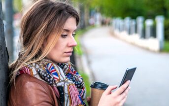 A woman with long hair, wearing a patterned scarf and brown jacket, stands outdoors holding a coffee cup and looking at her smartphone. Trees and a pathway are visible in the blurred background.
