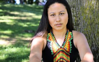 A woman with long dark hair sits outdoors by a tree, wearing a black top and a colorful beaded necklace with geometric patterns. Sunlight filters through the trees onto the grass in the background.