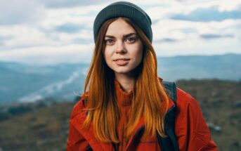A young woman with long red hair, wearing a gray beanie and a red jacket, stands outdoors with a blurred landscape of hills and cloudy sky in the background.