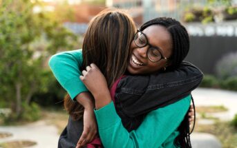 Two young women embrace outdoors, smiling warmly. One has long dark braids and glasses, while the other has straight brown hair. Greenery and buildings are blurred in the background.