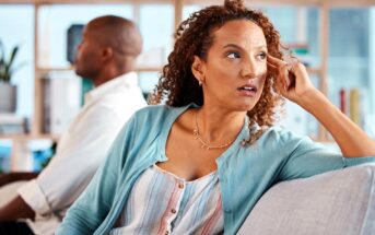 A woman with curly hair sits on a couch, looking away with a concerned expression, while a man sits in the background facing the opposite direction, both appearing upset or distant.