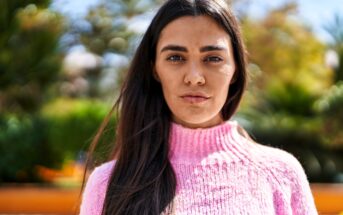 A woman with long dark hair wearing a pink sweater stands outdoors, looking directly at the camera. The background is blurred with green foliage and sunlight.
