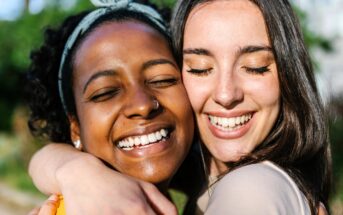 Two young women smiling with eyes closed, embracing each other outdoors on a sunny day, showing joy and friendship.
