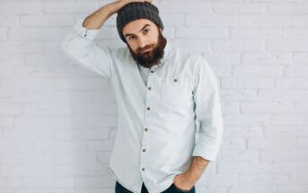 A bearded man in a gray knit beanie and light button-up shirt stands against a white brick wall, looking at the camera while holding one hand on top of his head.