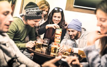 Five friends sit around a table with drinks, smiling and playing a game of Jenga. One woman is carefully removing a block while the others watch and laugh. The group appears relaxed and is warmly dressed.