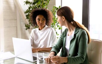 Two women sit at a desk in an office, having a conversation. One is wearing a white top and the other a green blazer. A laptop, papers, and a glass of water are on the desk, with plants and windows in the background.