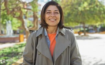 A woman with shoulder-length brown hair wearing a tan trench coat and orange top stands outdoors, smiling, with trees and sunlight in the background.
