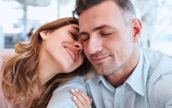 A smiling woman leans her head affectionately on a man’s shoulder. Both have their eyes closed, appearing content and relaxed. The background is softly blurred, suggesting a bright indoor setting.