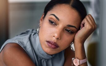 A woman with medium skin tone rests her face on her hand, looking thoughtfully into the distance. She wears a sleeveless gray top, silver hoop earrings, and a pink wristwatch.