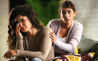 A woman sits on a couch, crying and wiping her face with a tissue, while another woman sits beside her, gently comforting her with a hand on her shoulder.