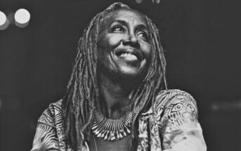Black and white photo of a smiling woman with long dreadlocks, wearing a patterned jacket and a large, spiked necklace, looking slightly upward against a dark background.
