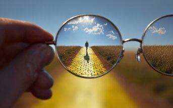 A hand holds glasses that focus on a person walking down a vivid yellow brick road through a field, making the road appear sharp and clear inside the lenses, while the outside is blurred.