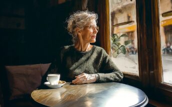 An older woman with gray curly hair and glasses sits at a round table by a window, holding a cup of coffee and gazing outside. Warm sunlight illuminates her face and the cozy café interior.