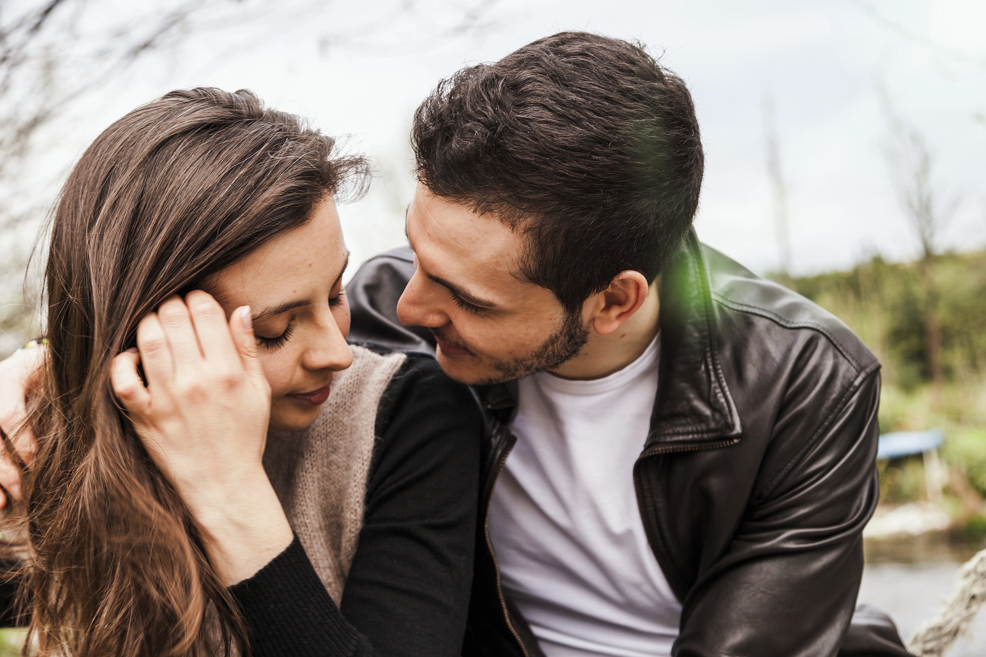 A couple sitting close together outdoors. The man is wearing a leather jacket and white shirt, looking lovingly at the woman, who is smiling with her head slightly turned. She has long brown hair and a relaxed expression. Trees and grass are in the background.