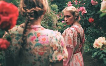 Two young women with braided hair and floral dresses stand among blooming roses. One faces away, while the other looks back over her shoulder with a serious expression. The scene is lush and colorful.