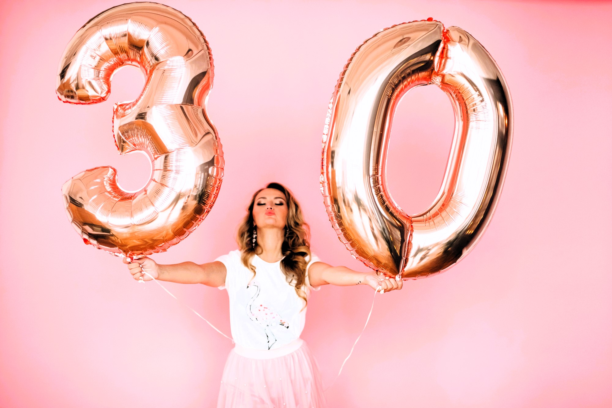 Woman in a white shirt and pink skirt holds large rose gold balloons shaped as the number "30" against a pink background. She is puckering her lips and standing with arms outstretched.
