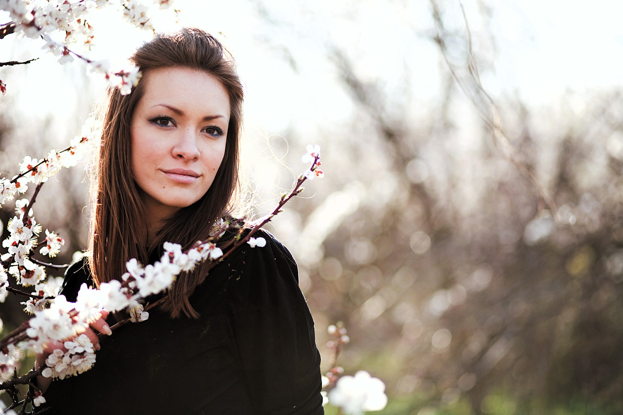 A woman with long brown hair stands near a branch of blooming flowers. She wears a black top and looks towards the camera with a gentle expression. The background is softly blurred, highlighting the bright blossoms.
