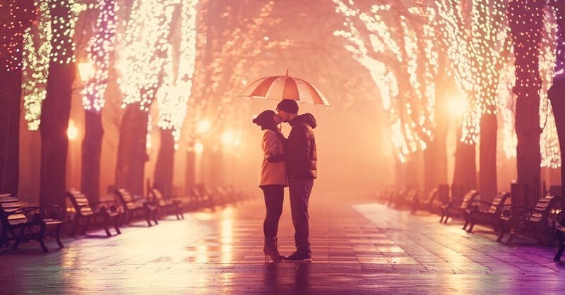 couple kissing under umbrella down a boulevard with lights