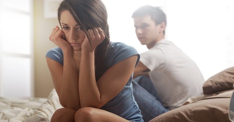 young couple sitting on bed facing apart, the woman looking disappointed