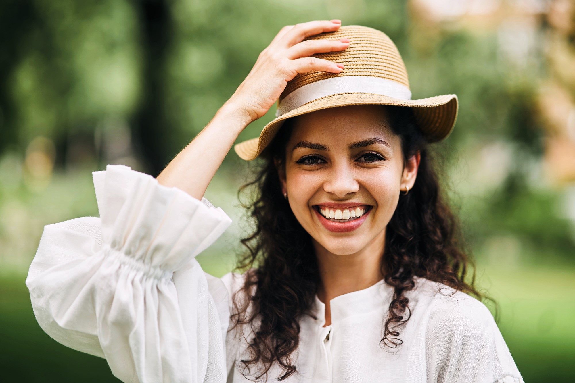 A woman with curly hair smiles brightly, wearing a straw hat and a white blouse. She is outdoors with a blurred green background. Her hand is gently touching the hat, and she appears joyful and relaxed.