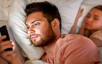 man having emotional affair by texting someone else whilst girlfriend sleeps in the background