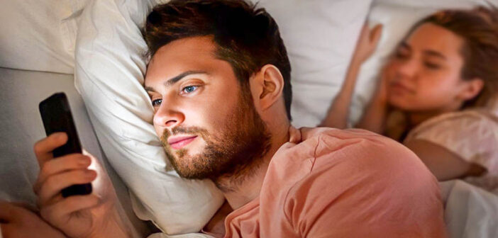 man having emotional affair by texting someone else whilst girlfriend sleeps in the background