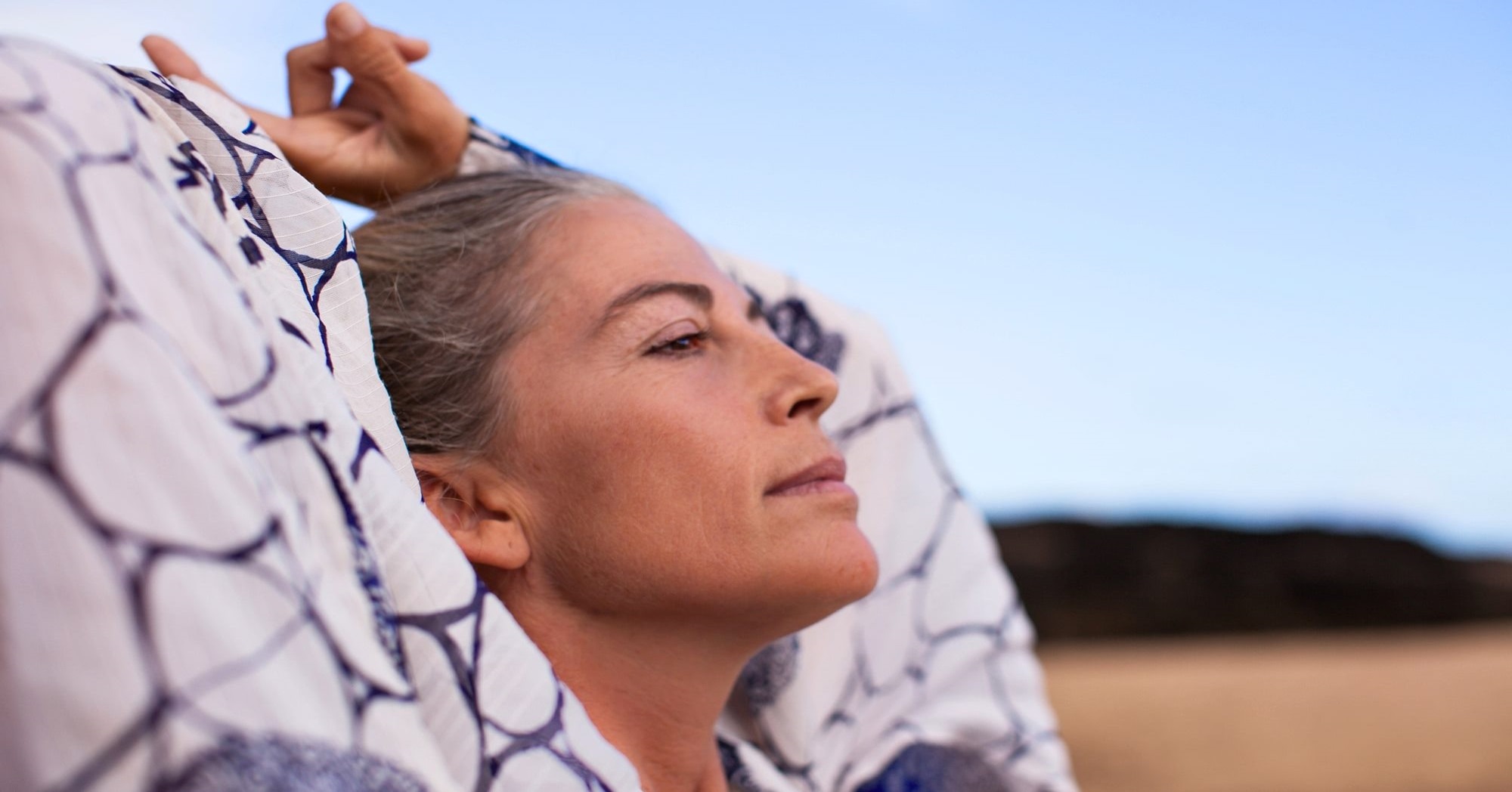 A person with light skin and gray hair relaxes outdoors with arms raised behind their head, wrapped in a white fabric with blue patterns. They appear calm and focused, gazing upward against the backdrop of a clear blue sky and distant landscape.