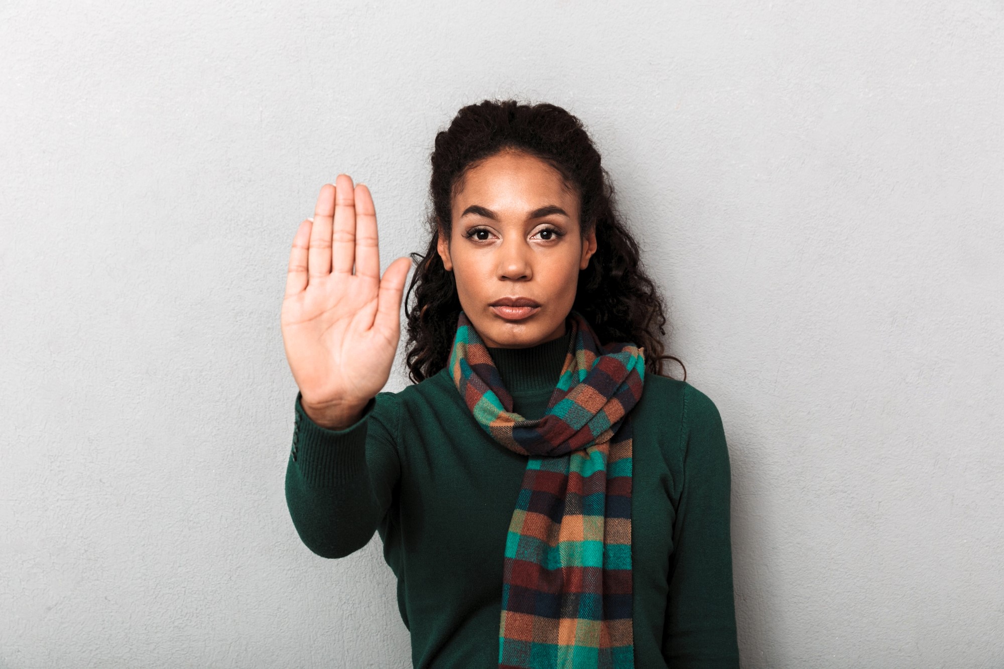 A woman with long, dark curly hair stands against a gray background. She is wearing a green sweater and a multicolored scarf. Her right hand is raised with her palm facing outward, conveying a gesture to stop or halt.