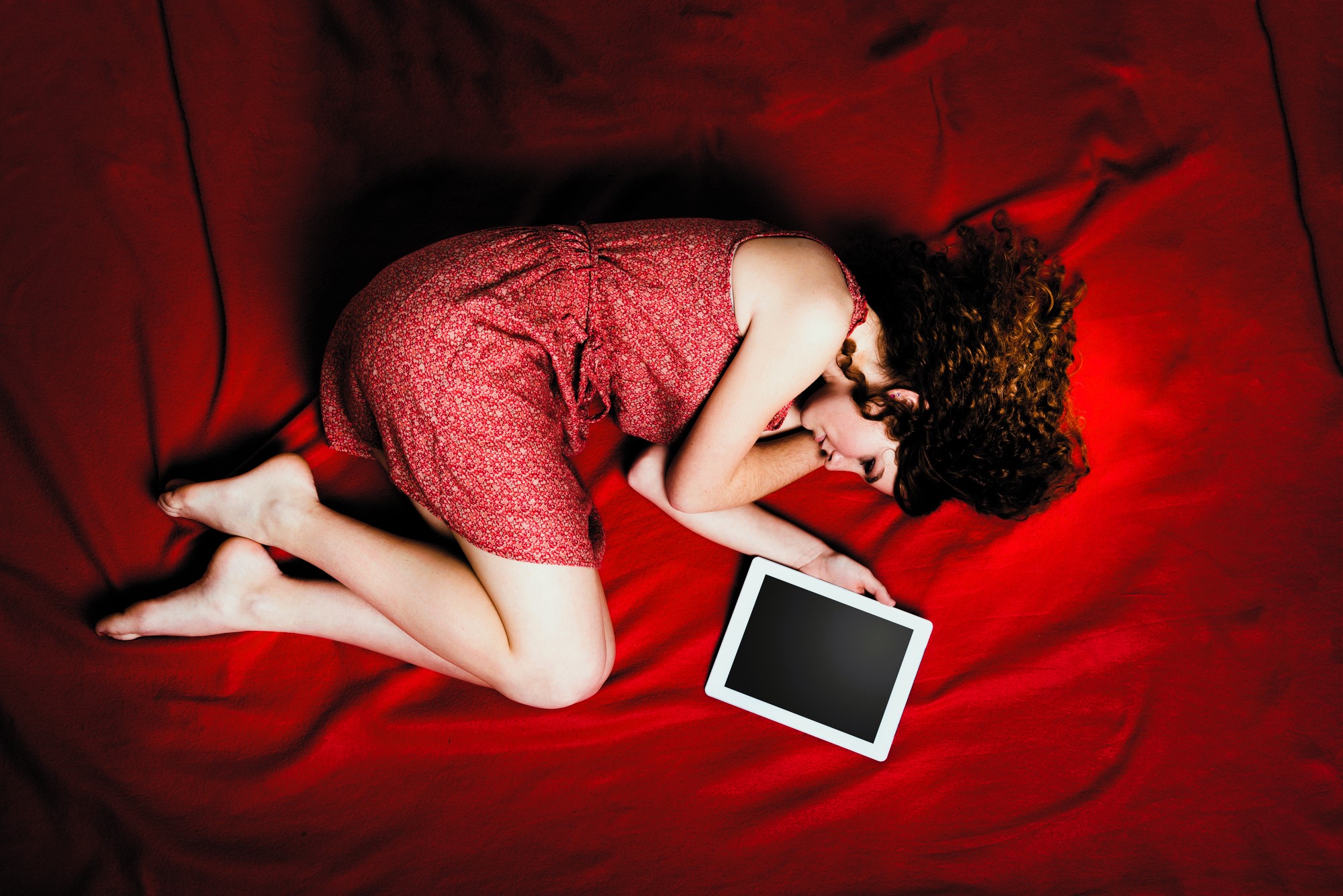 A woman with curly hair lies on a red blanket, wearing a red dress. She is curled up on her side with her head resting on one arm and is holding a tablet with a blank screen in her other hand.