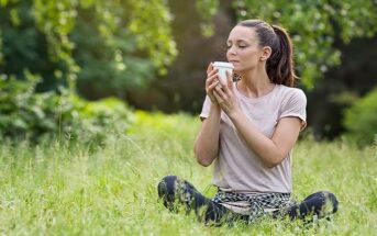 a woman sitting on grass holding and smelling a takeaway coffee - illustrating the 54321 grounding technique