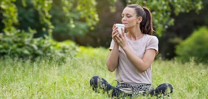 a woman sitting on grass holding and smelling a takeaway coffee - illustrating the 54321 grounding technique