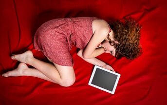 sad woman curled up on bed next to tablet signifying a long-distance breakup