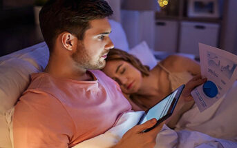 workaholic husband looking at tablet computer and paperwork in bed whilst his wife sleeps